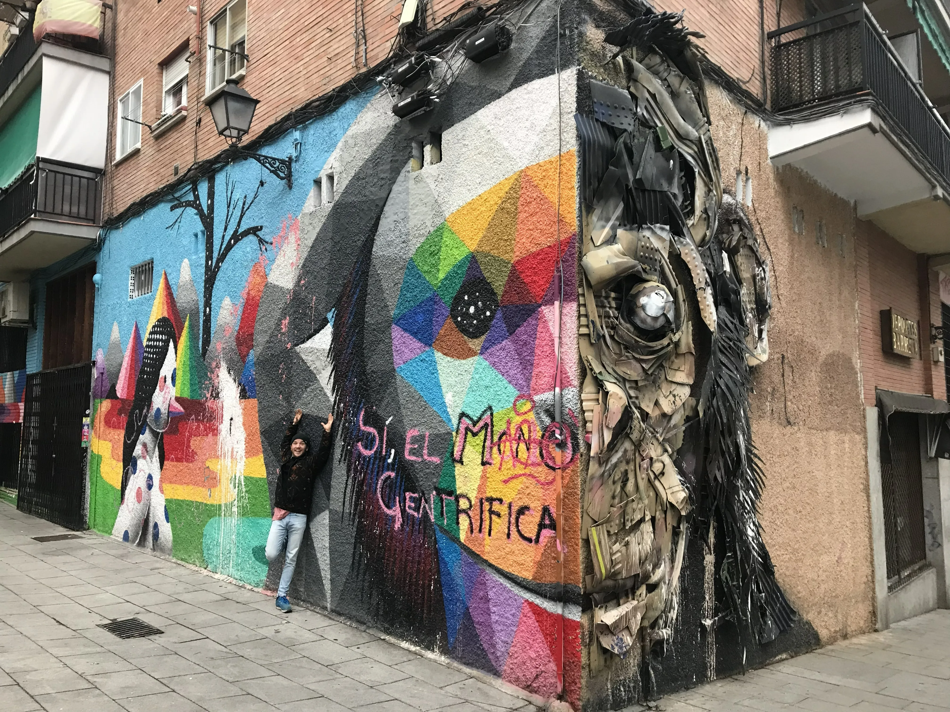 Fresque murale monumentale à l’angle d’un immeuble : visage géométrique multicolore et relief sculpté en matériaux de récupération, un passant bras levés au pied du mur donne l’échelle
