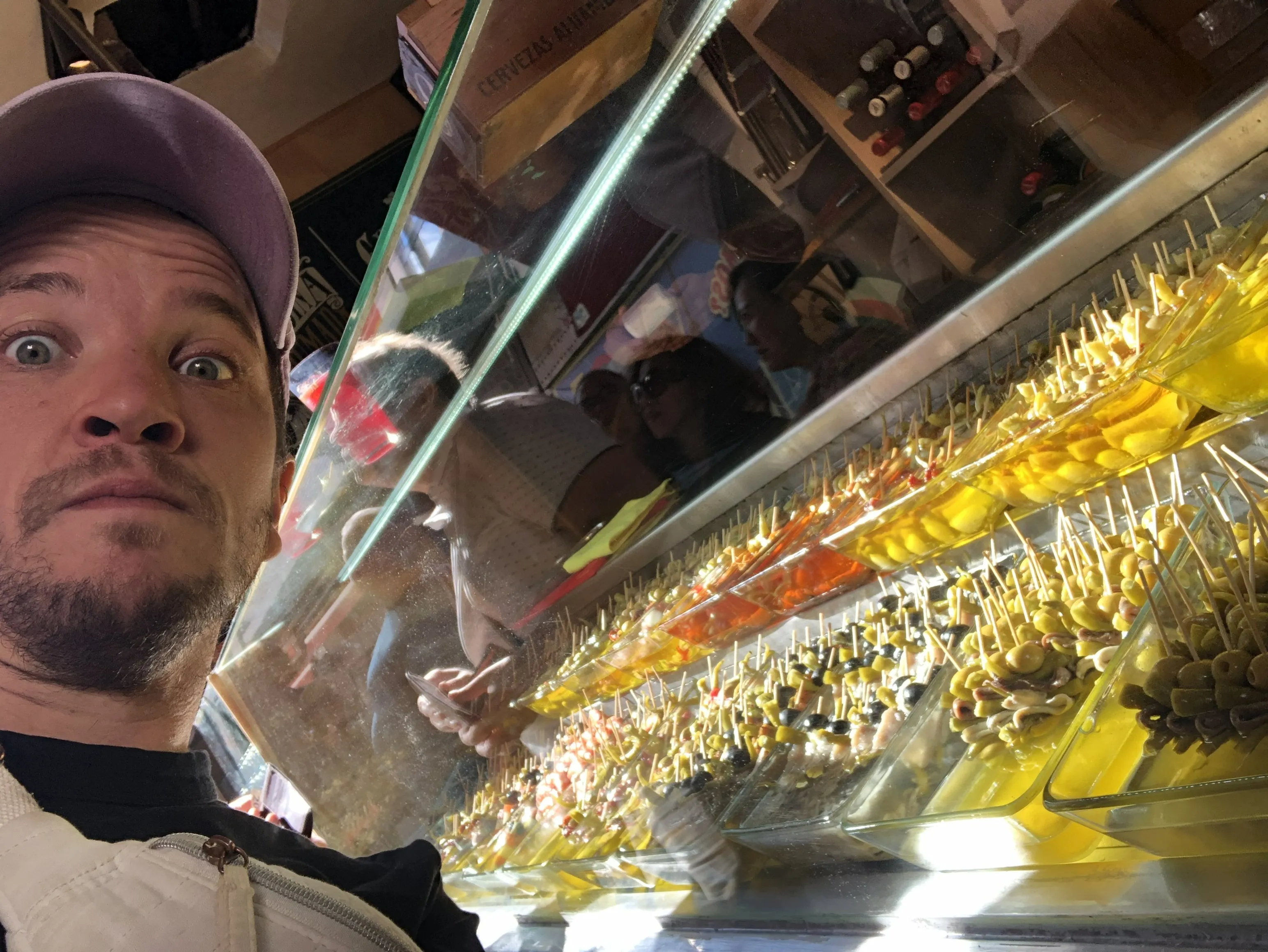 Lucas, casquette violette, au premier plan devant la vitrine d'un bar à tapas : rangées de banderilles, olives et cornichons sur pique, clients attablés en arrière-plan