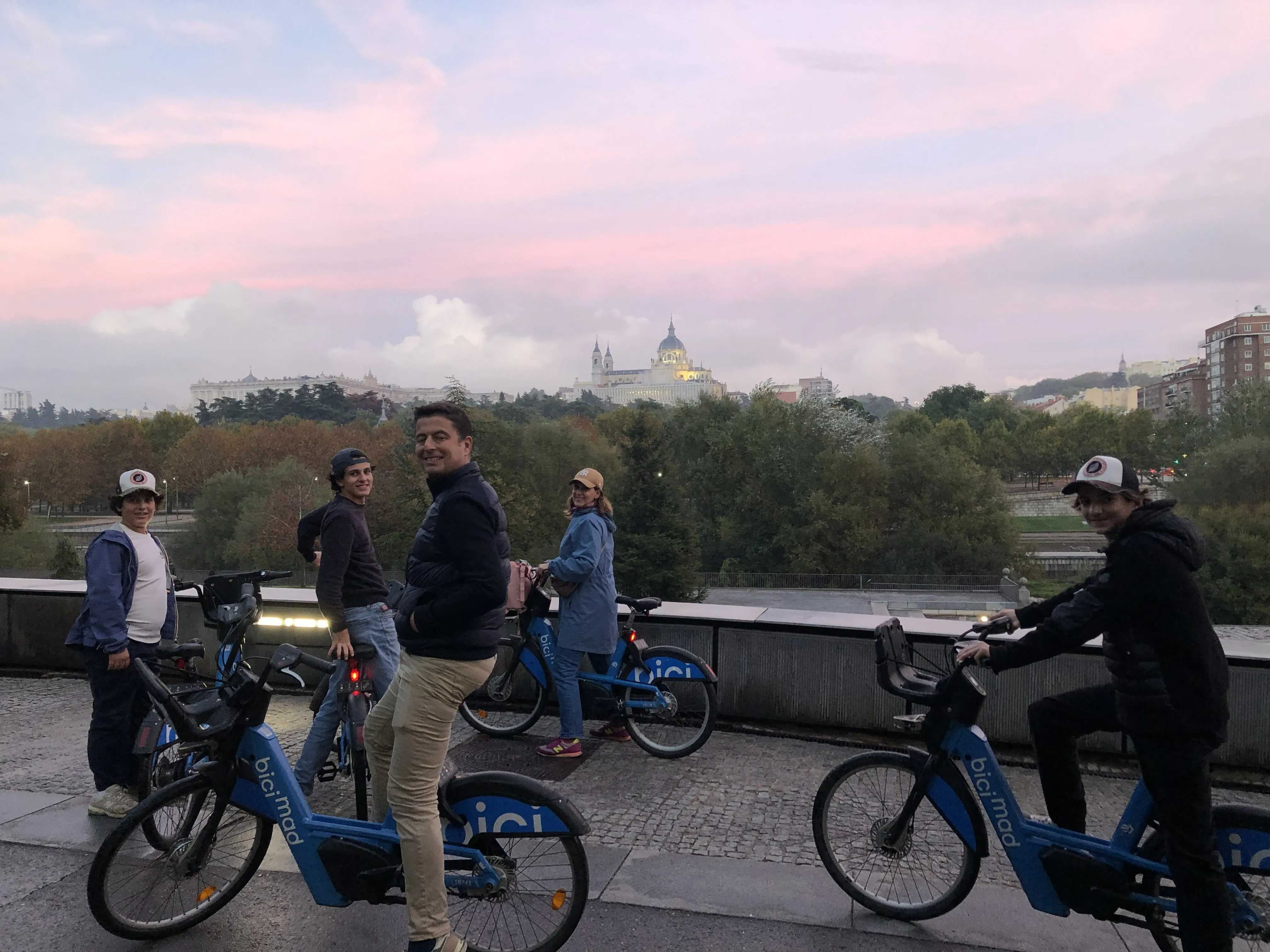 Un groupe familial à vélo électrique BiciMAD à la tombée du jour, ciel rose, le Palais royal et la cathédrale de l’Almudena au loin
