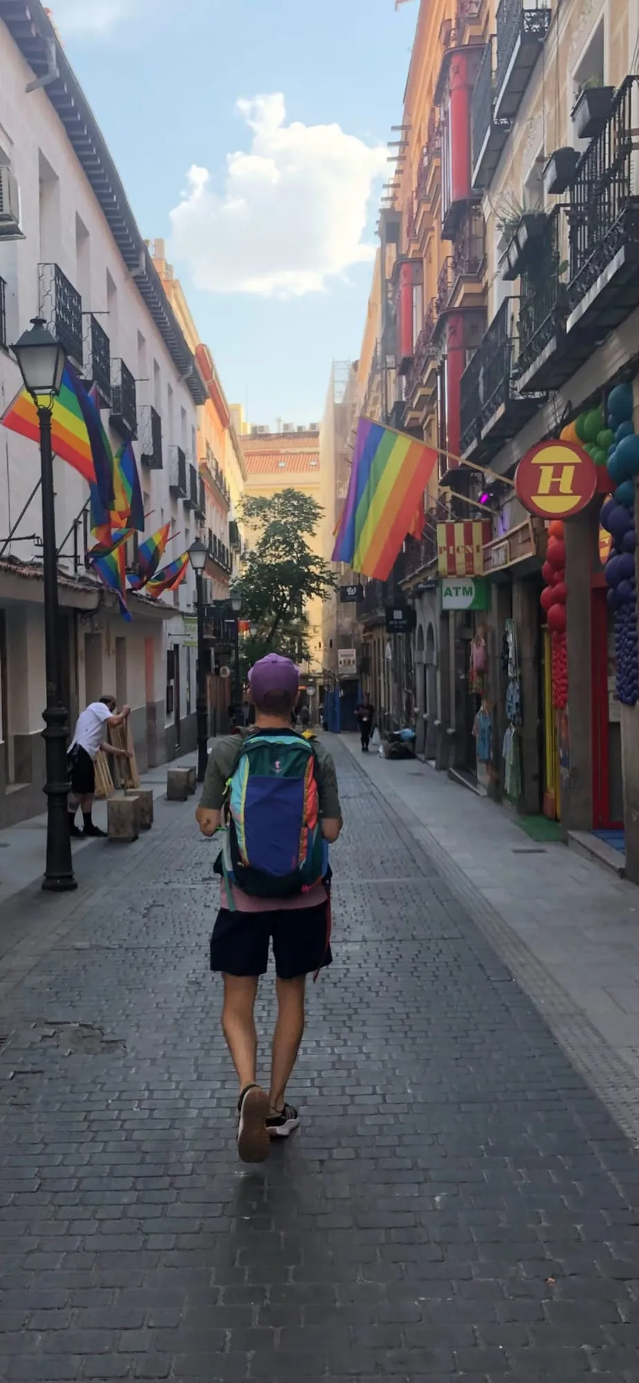 Lucas de dos, casquette violette et sac à dos coloré, marchant dans une rue pavée de Chueca pavoisée de drapeaux arc-en-ciel