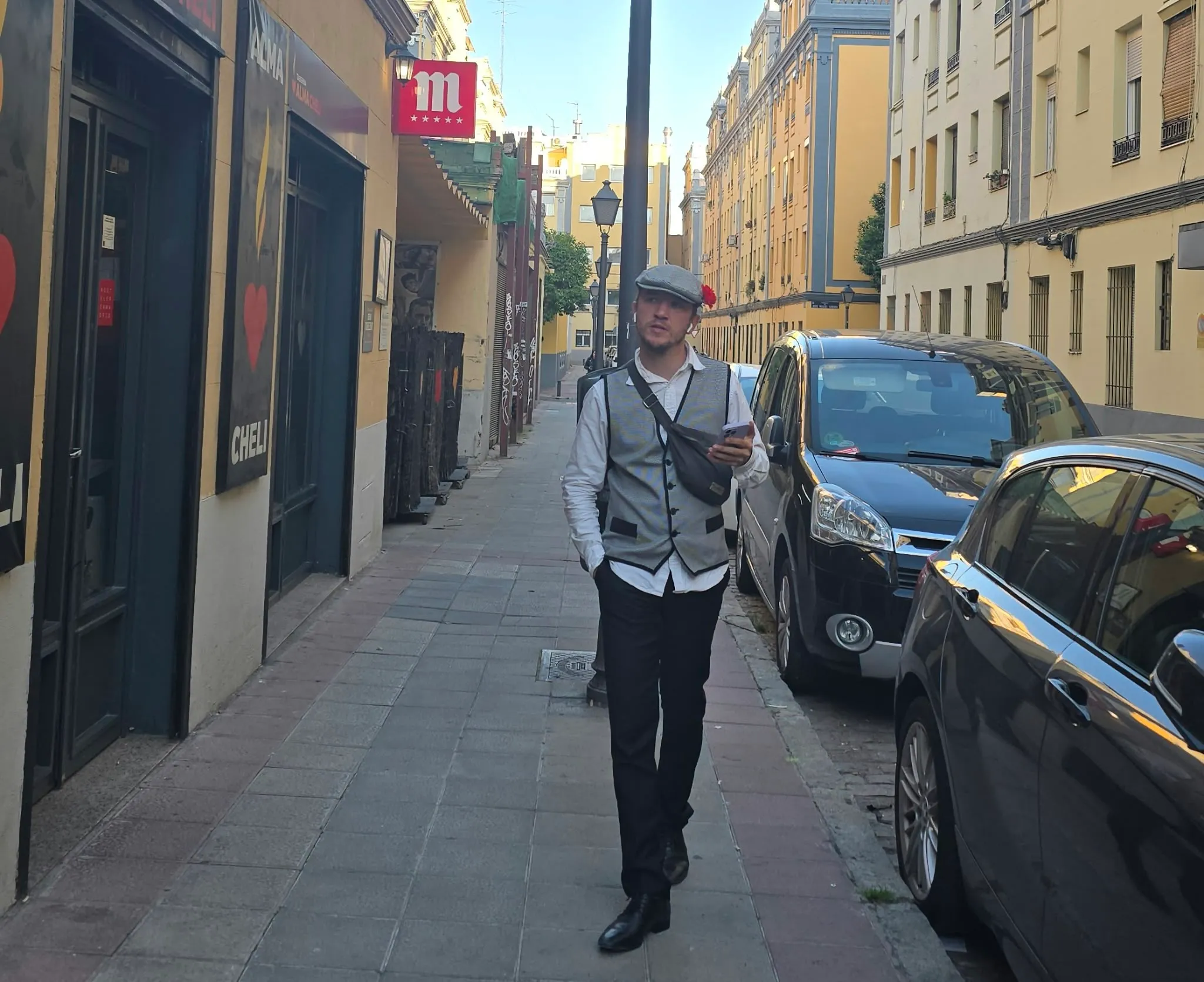 Lucas, casquette plate et œillet rouge, gilet et sacoche en bandoulière, marche téléphone à la main dans une rue de Madrid bordée de voitures garées