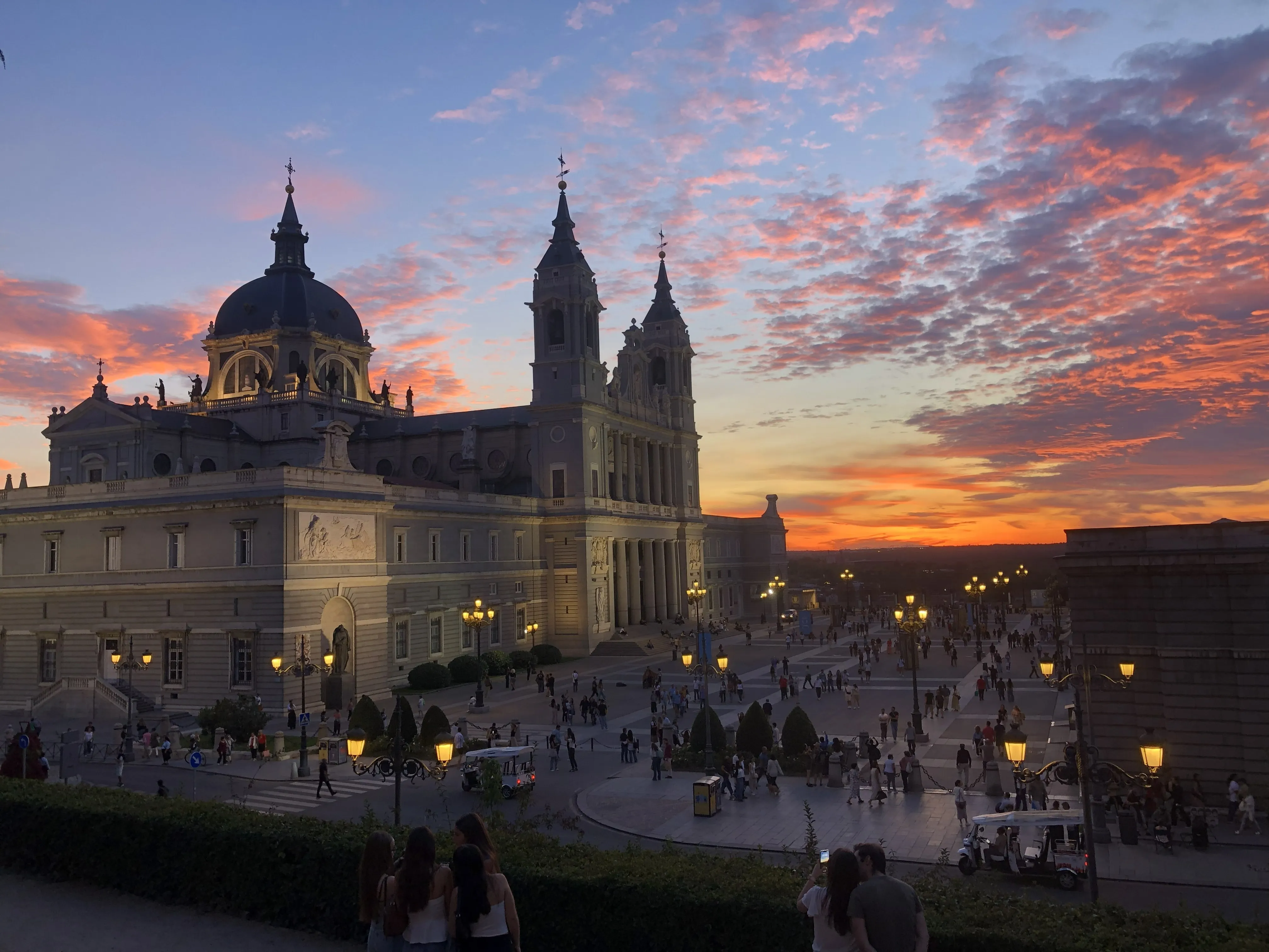 La cathédrale de l’Almudena au coucher du soleil, ciel orange strié de nuages, promeneurs sur l’esplanade et badauds attablés au premier plan
