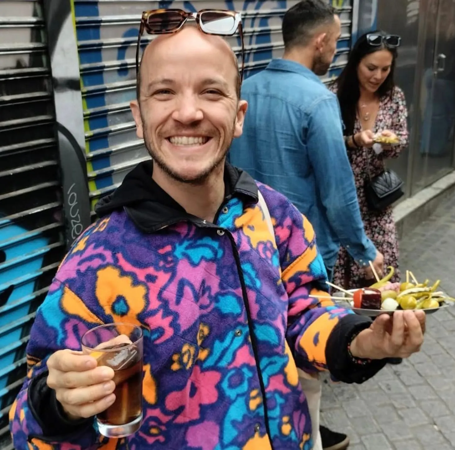 Lucas souriant, une assiette de tapas dans une main et un vermouth dans l'autre, devant un rideau de fer graffé à Lavapiés