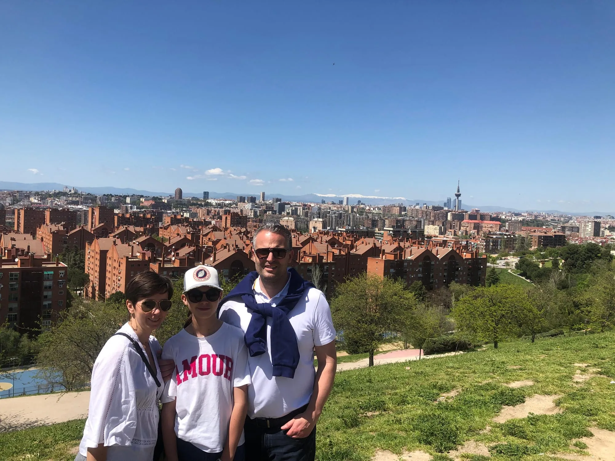 Une famille pose en haut d'une colline herbeuse, avec derrière elle tout Madrid en panorama, la tour Torrespaña et les sommets enneigés de la sierra de Guadarrama