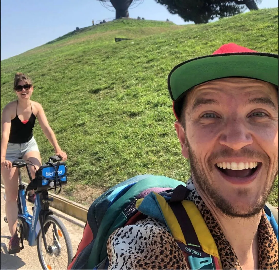 Lucas en casquette et une visiteuse à vélo électrique BiciMAD sur une allée bordée de pelouse dans un parc de Madrid