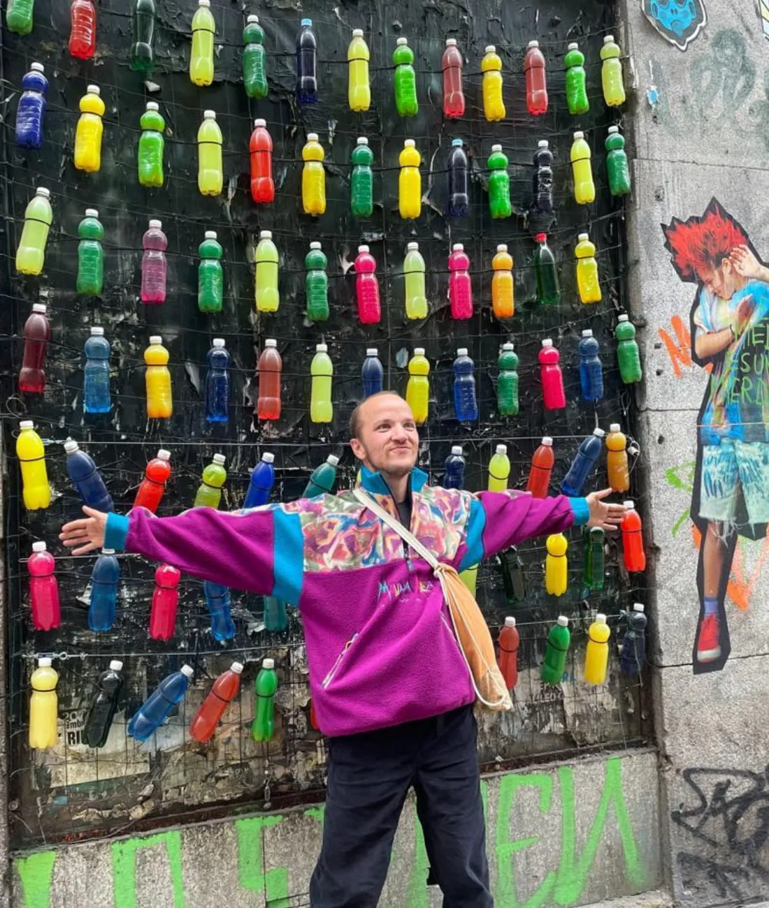 Lucas bras écartés devant un mur couvert de bouteilles en plastique colorées, œuvre de street art dans une rue de Madrid
