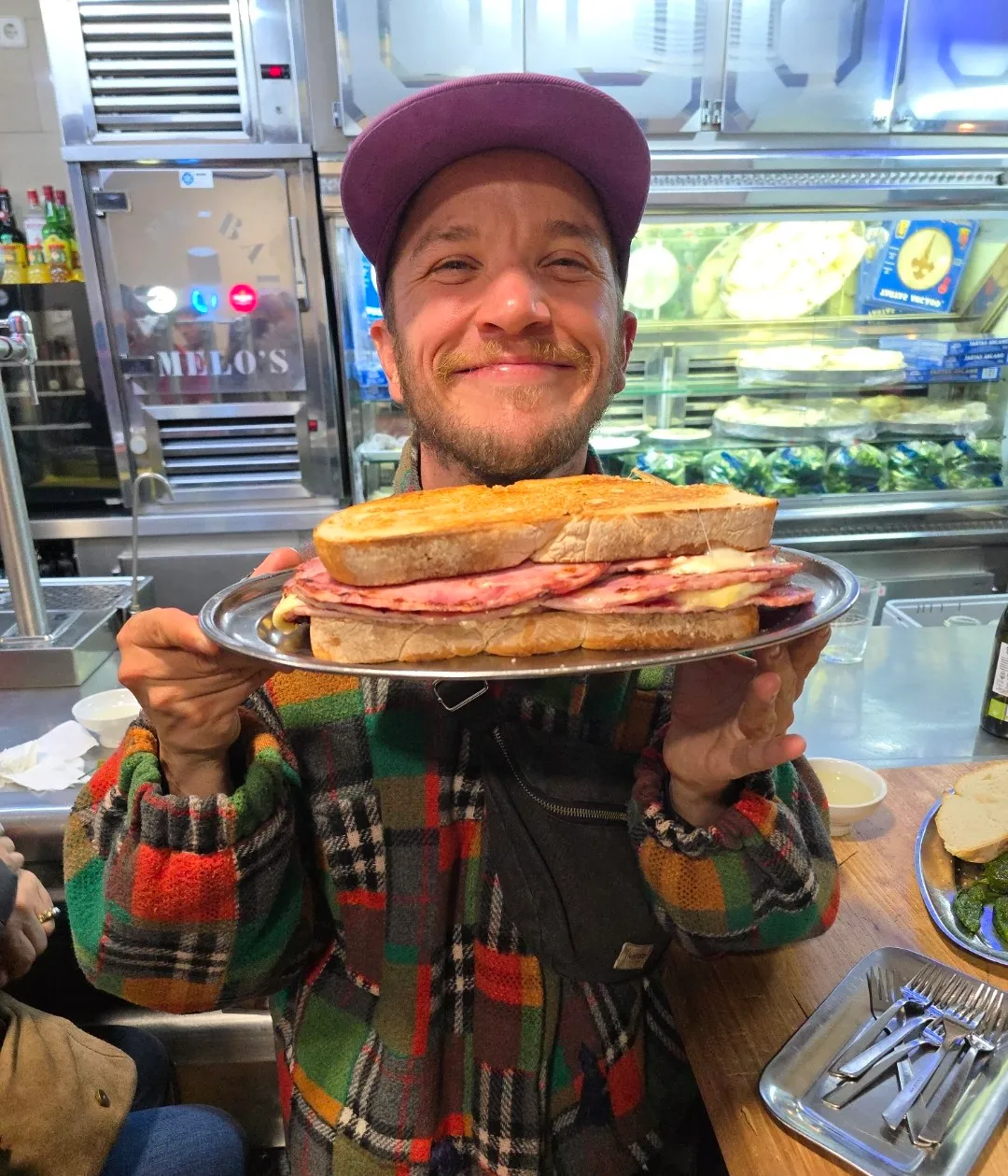 Lucas, casquette violette et veste à carreaux colorée, présente en souriant un immense sandwich au jambon et au fromage sur un plateau, au comptoir du bar Melo's
