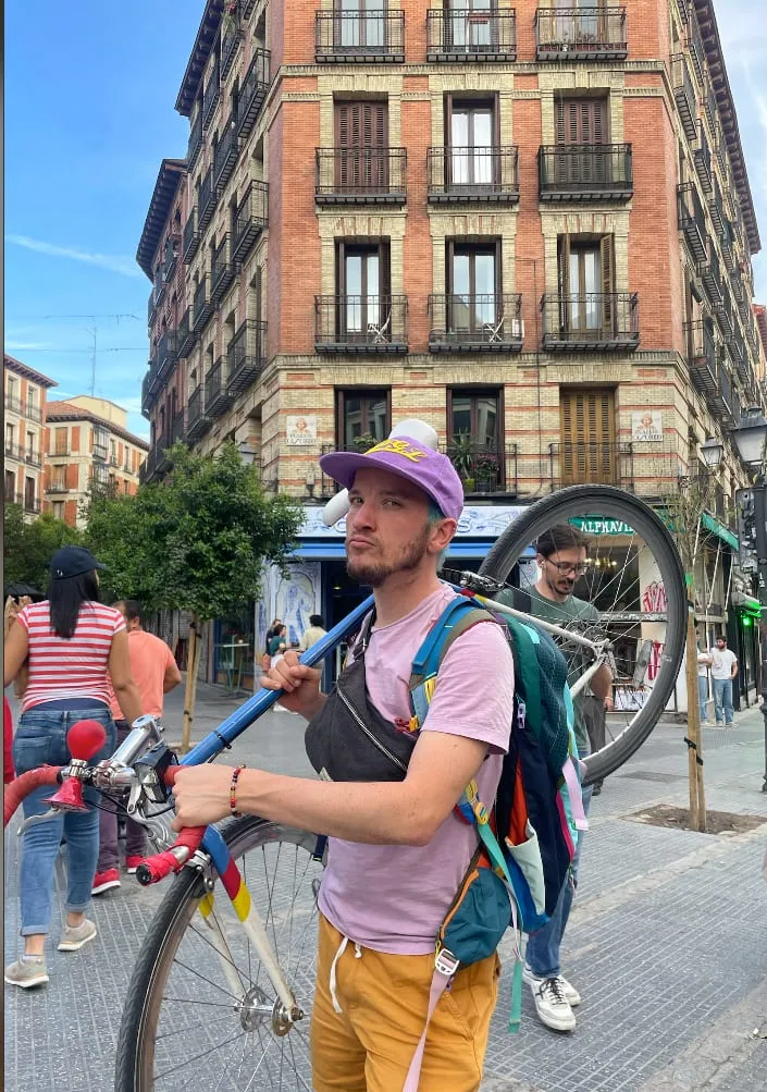 Lucas portant un vélo sur l'épaule dans une rue de Madrid, immeuble en brique rouge en arrière-plan